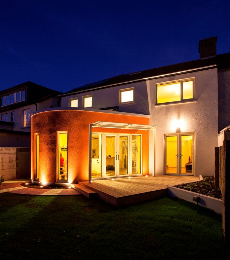 Image of the EnerPHit project completed by Joseph Little Architects and Anne-Marie Fallon. Two-story dwelling with many windows and a round orange extension on the first floor.